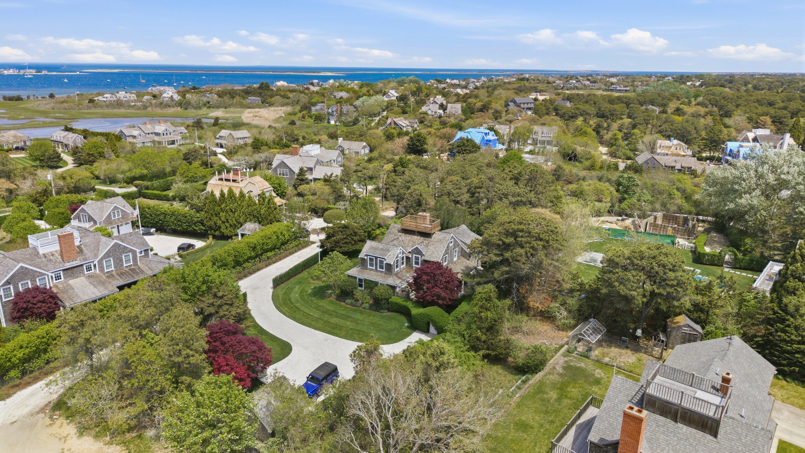 Aerial view of a shingled home with a rooftop deck, landscaped grounds, and harbor in the distance.