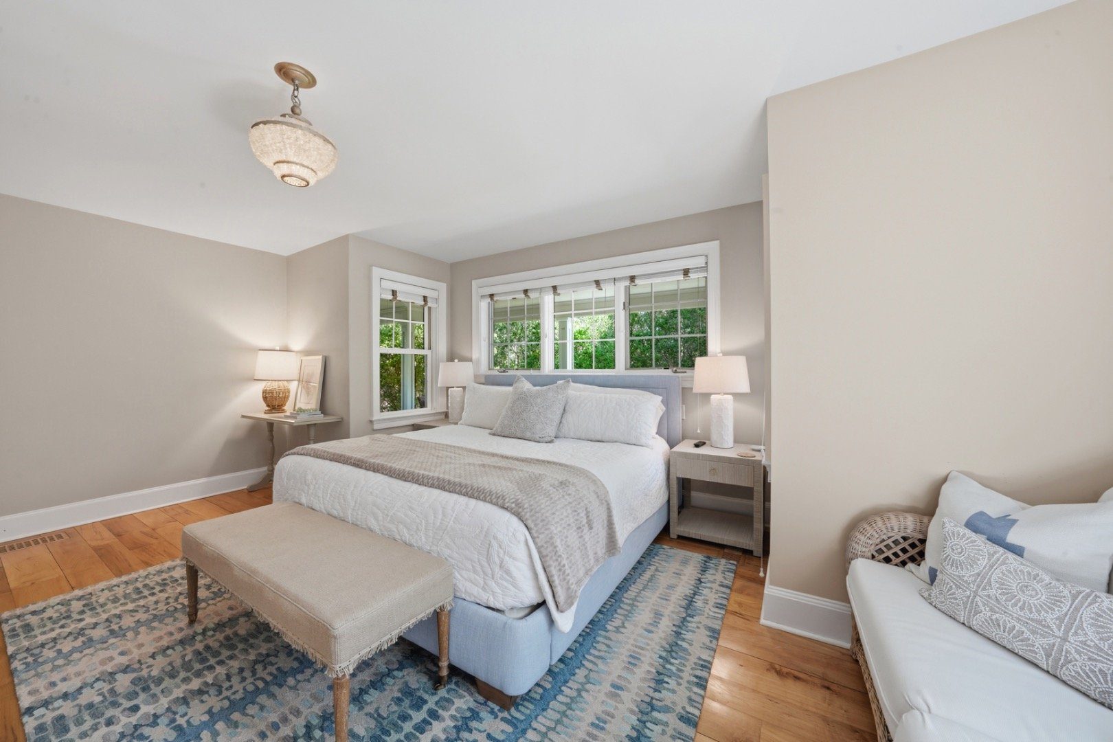 Bedroom with a white quilted bed, blue patterned rugs, and windows overlooking greenery.