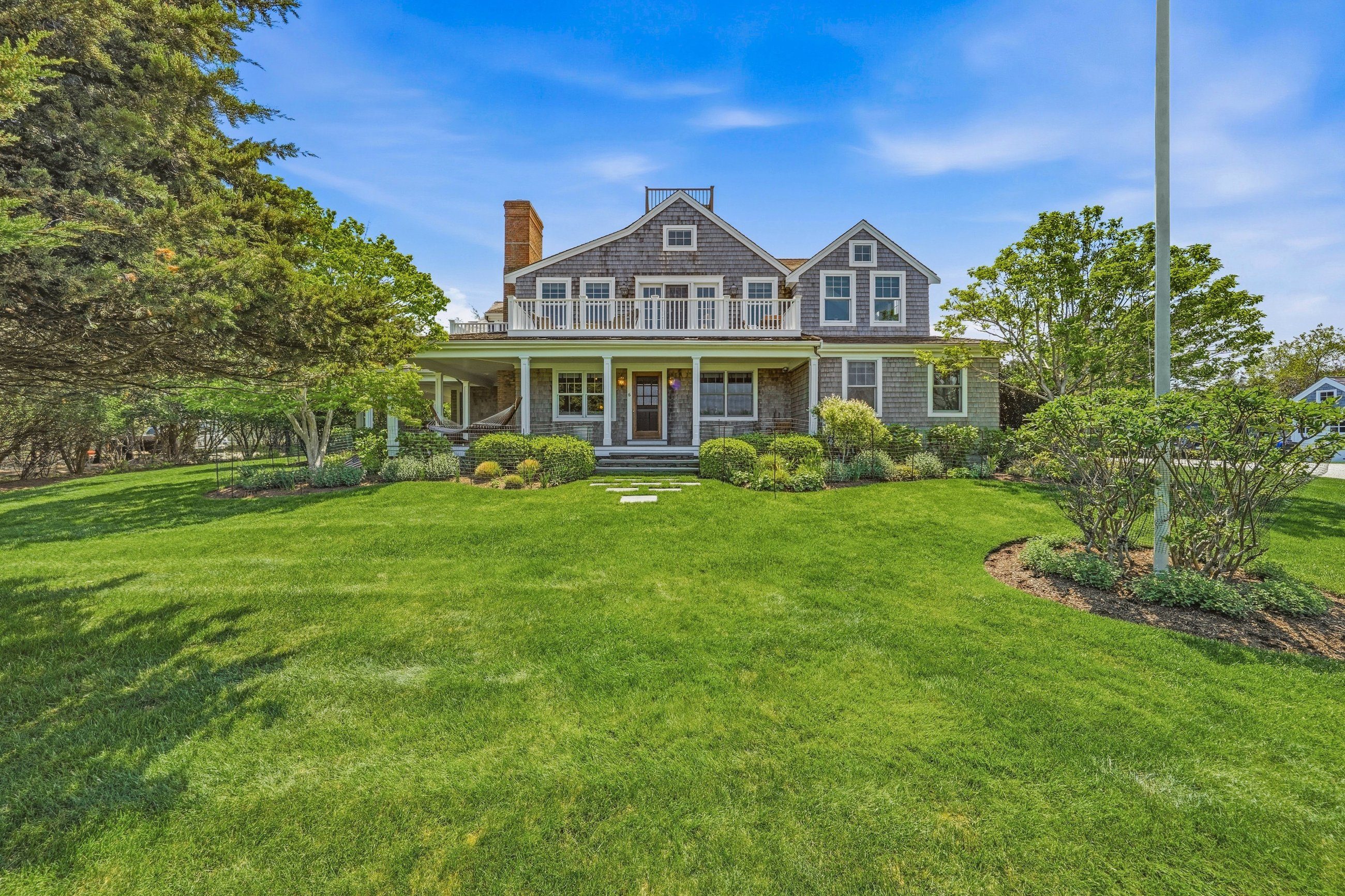 Gray-shingled home with a covered porch and upper balcony beyond a broad green lawn.