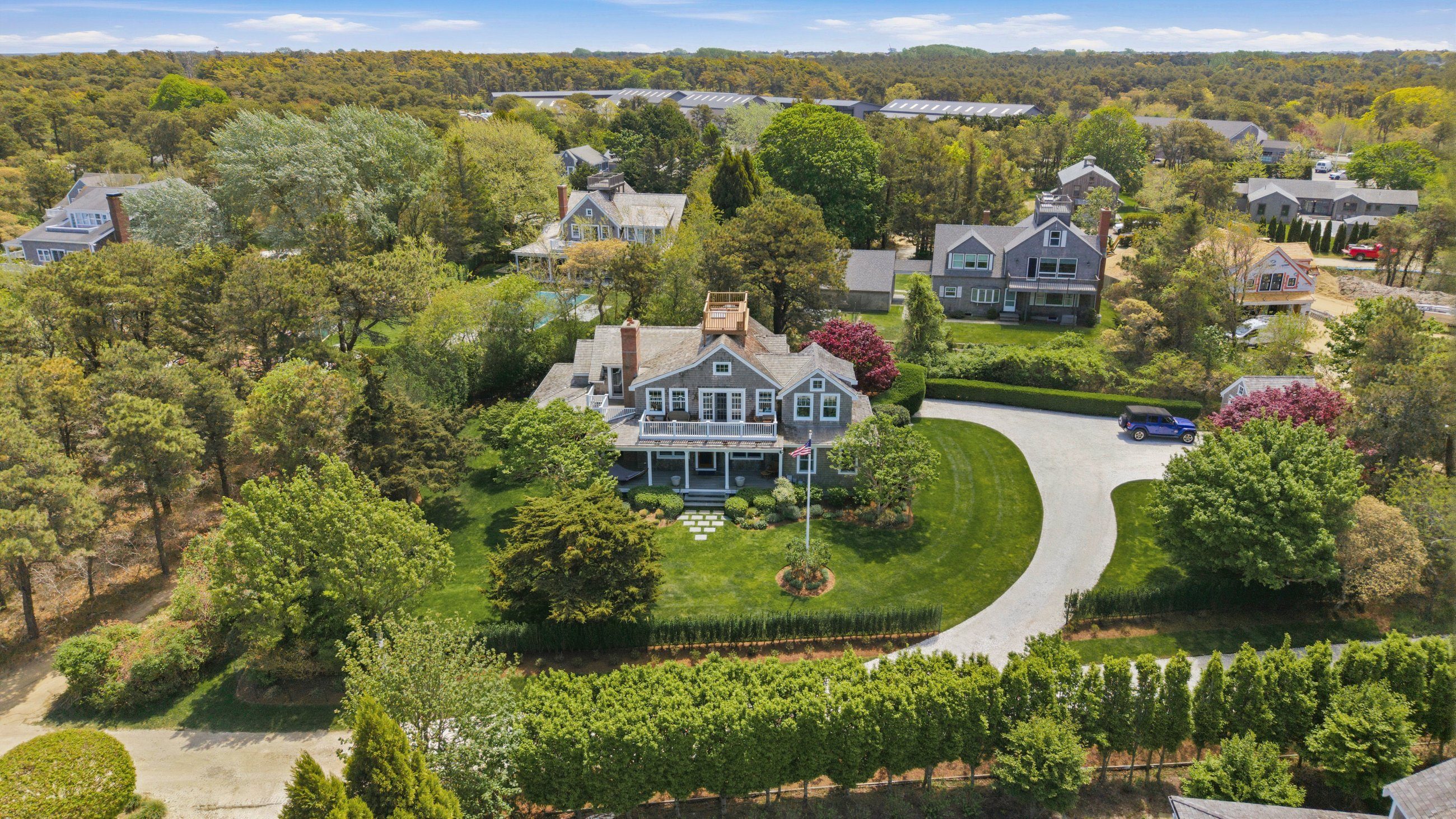 Aerial view of a shingled Nantucket home with a curved driveway, landscaped lawn, and surrounding trees.