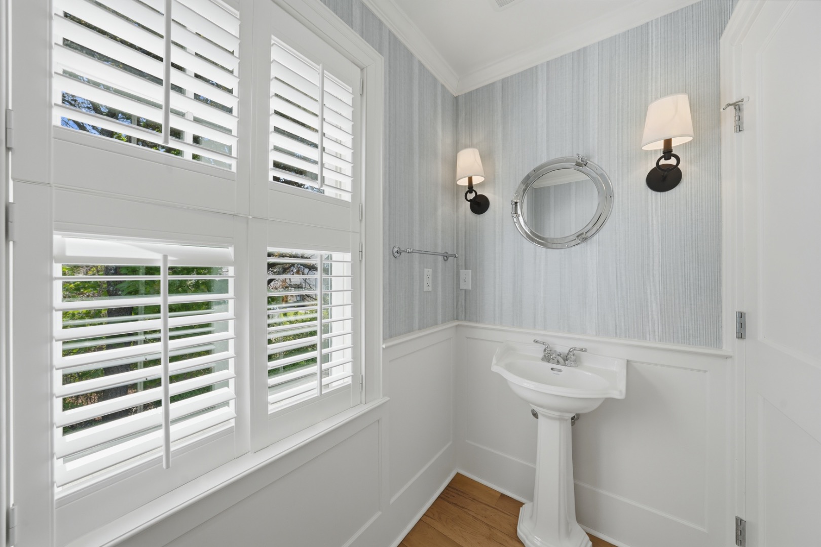 Bathroom with a pedestal sink, oval chrome-framed mirror, and plantation shutters.