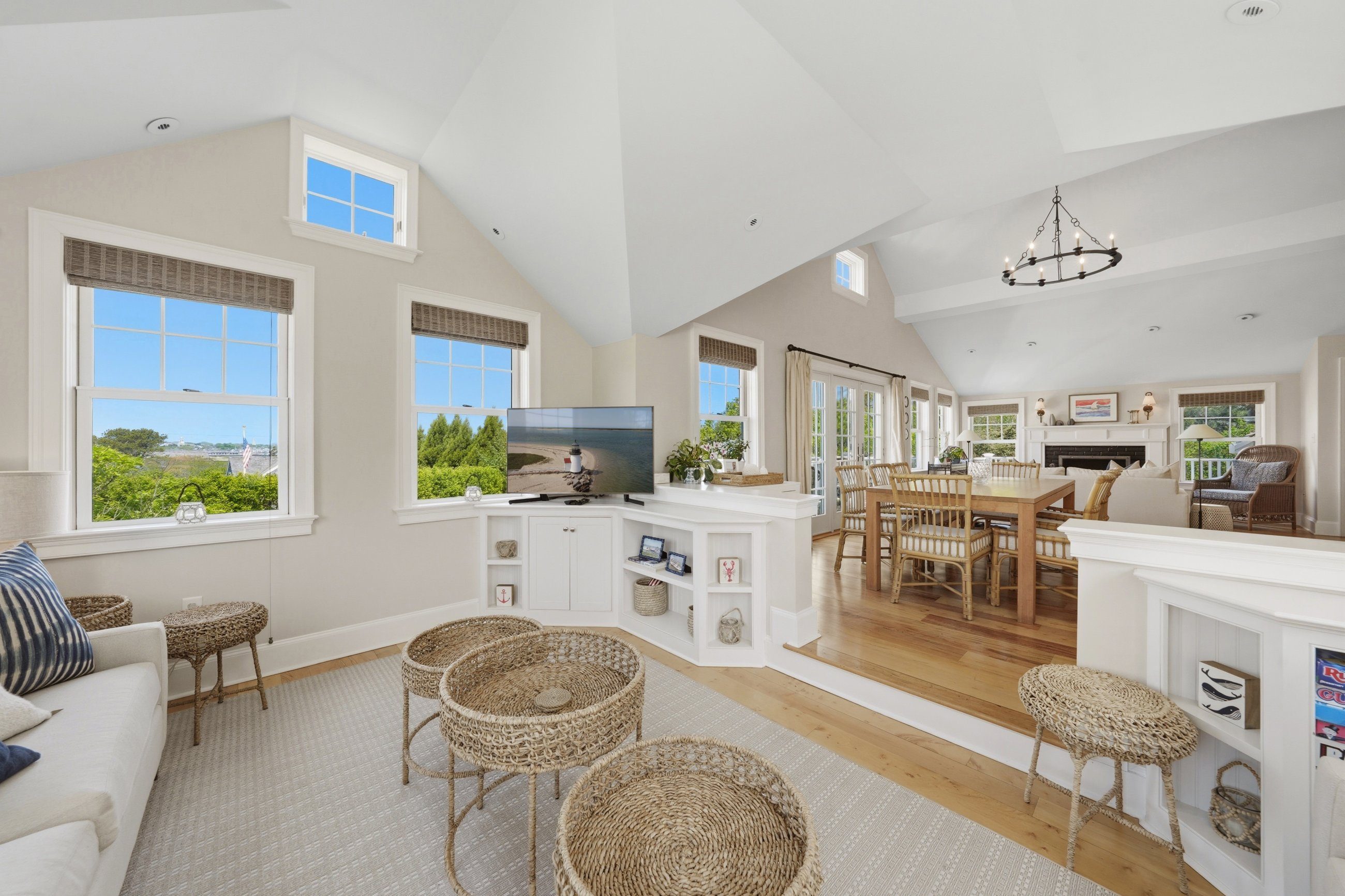 Open-plan living and dining room with a vaulted ceiling, wicker furniture, white built-ins, and large windows.
