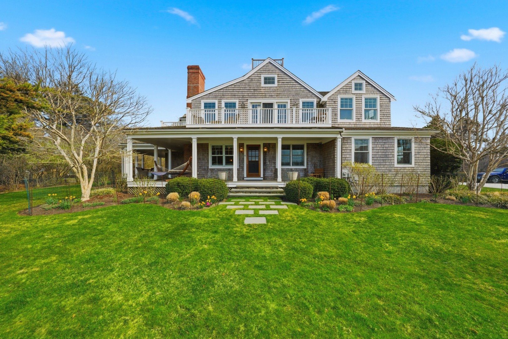 Shingle-style house with a covered porch and upper balcony, fronted by a landscaped garden and green lawn.