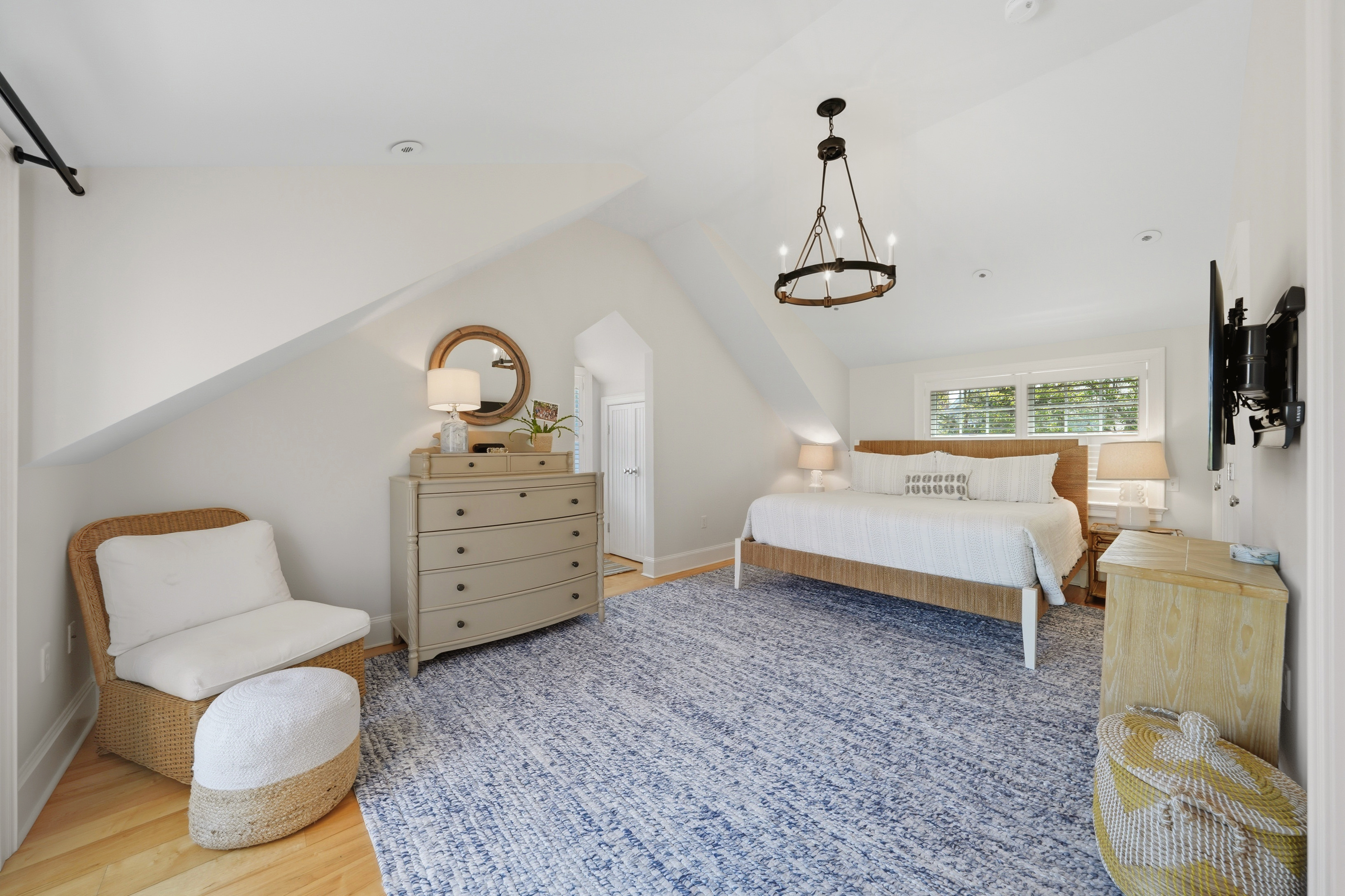 Furnished bedroom with a white bed, blue-gray rug, wicker chair, and sloped ceiling.