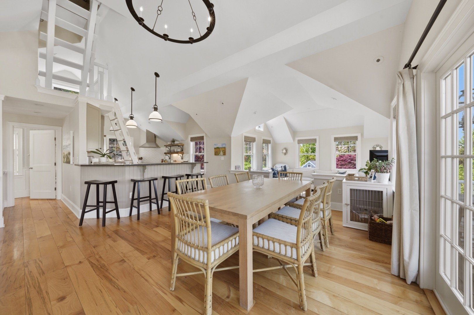Dining room with a long wood table, woven chairs, vaulted ceiling, and adjoining kitchen.