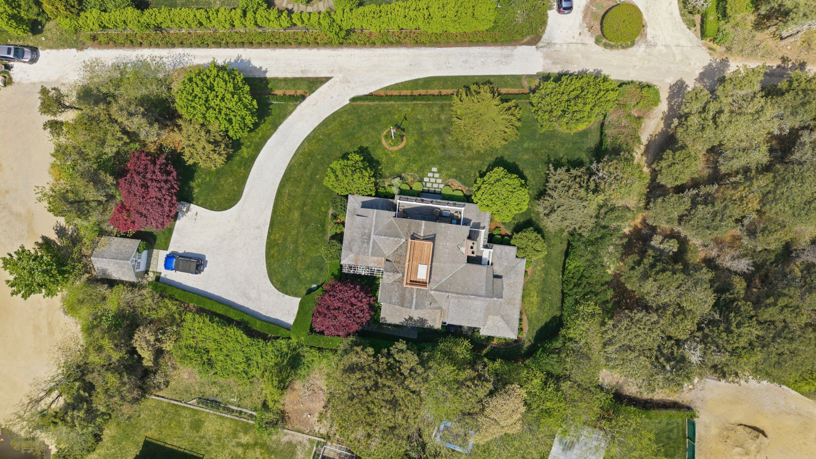 Aerial view of a house amid mature trees, with a curved gravel driveway and landscaped grounds.