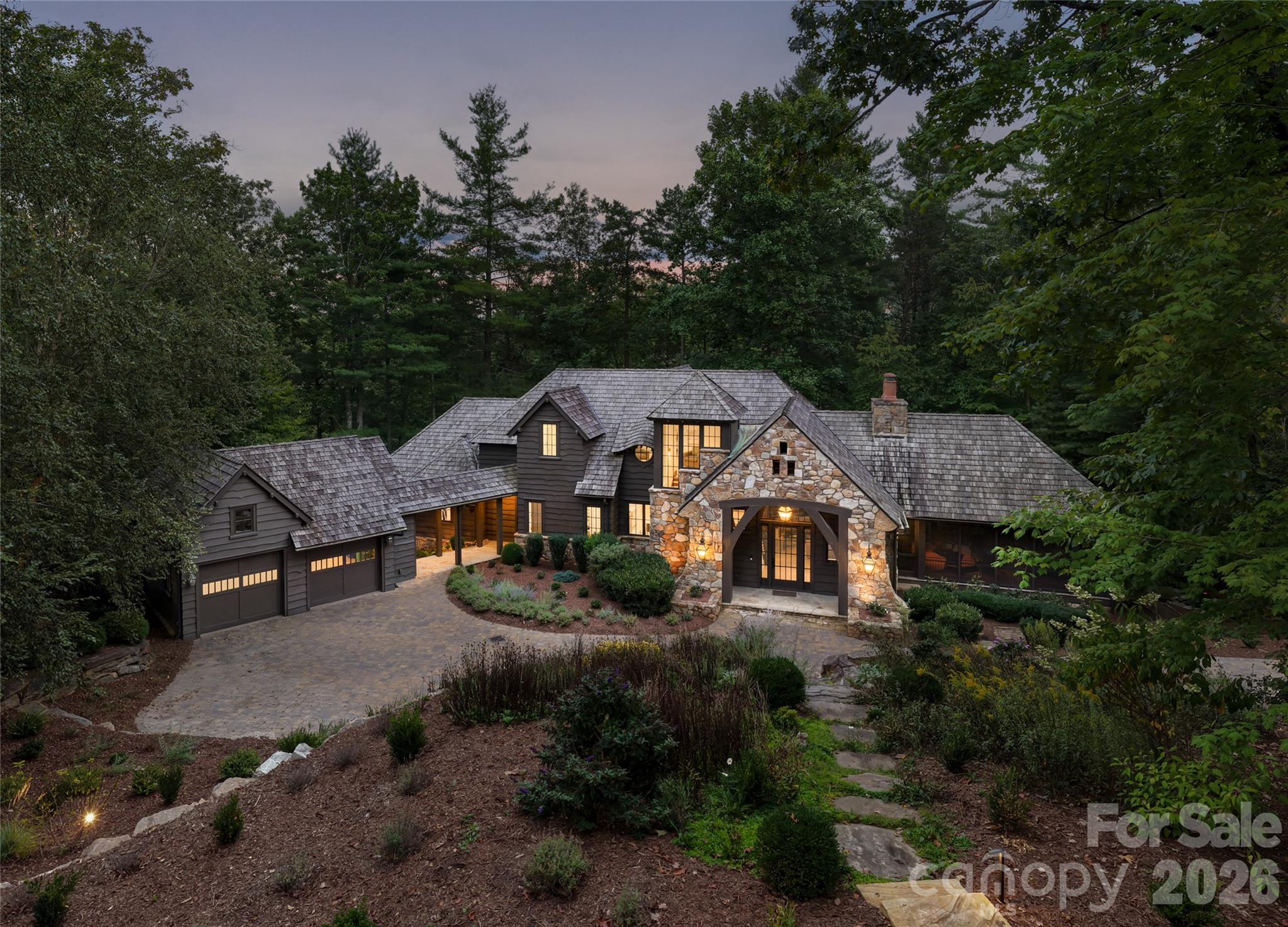 Wooded home with a stone entryway, illuminated windows, landscaped grounds, and a broad paved driveway at dusk.