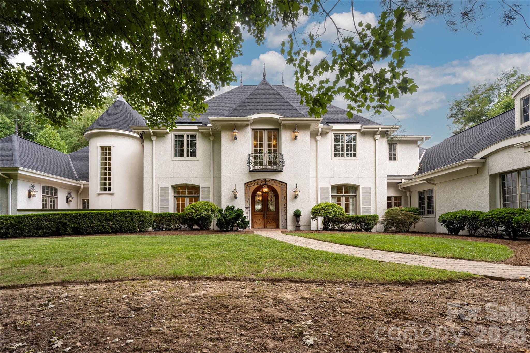White stucco home with a round turret, central balcony, and arched entrance behind a stone walkway and lawn.