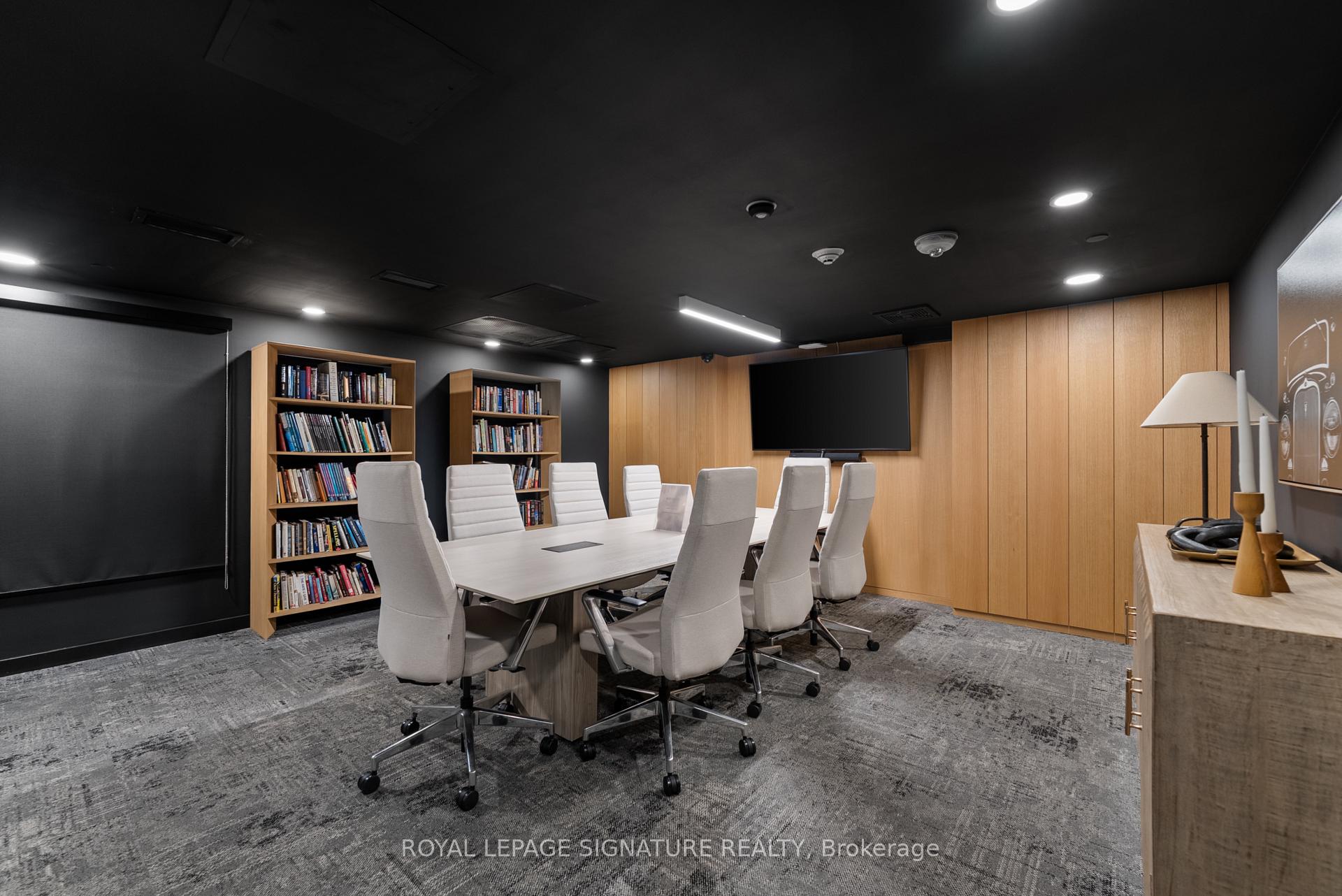 Conference room with a long meeting table, white chairs, bookshelves, and a wall-mounted screen.
