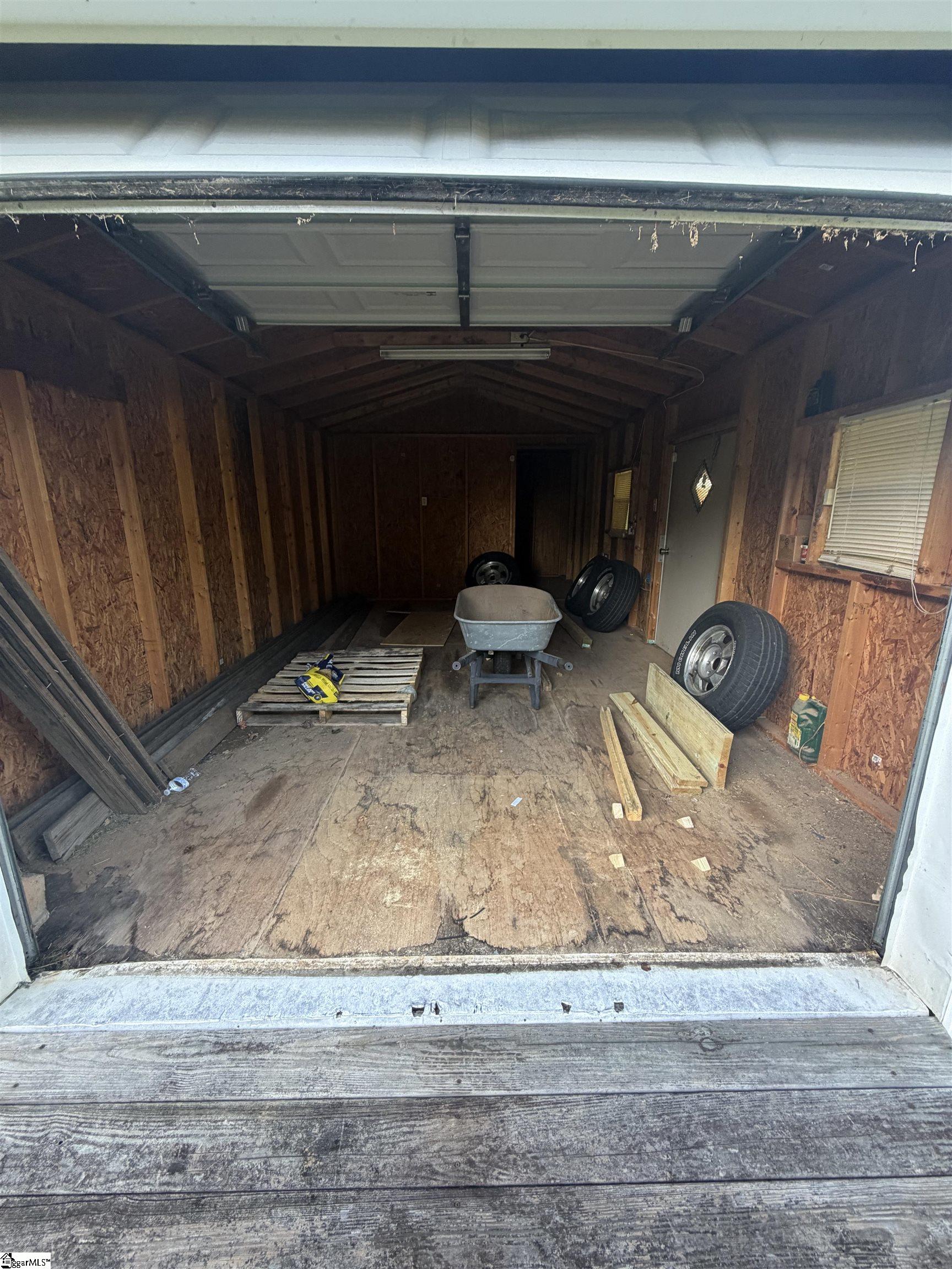 Wooden garage interior with an open overhead door, tires, lumber, and a wheelbarrow.