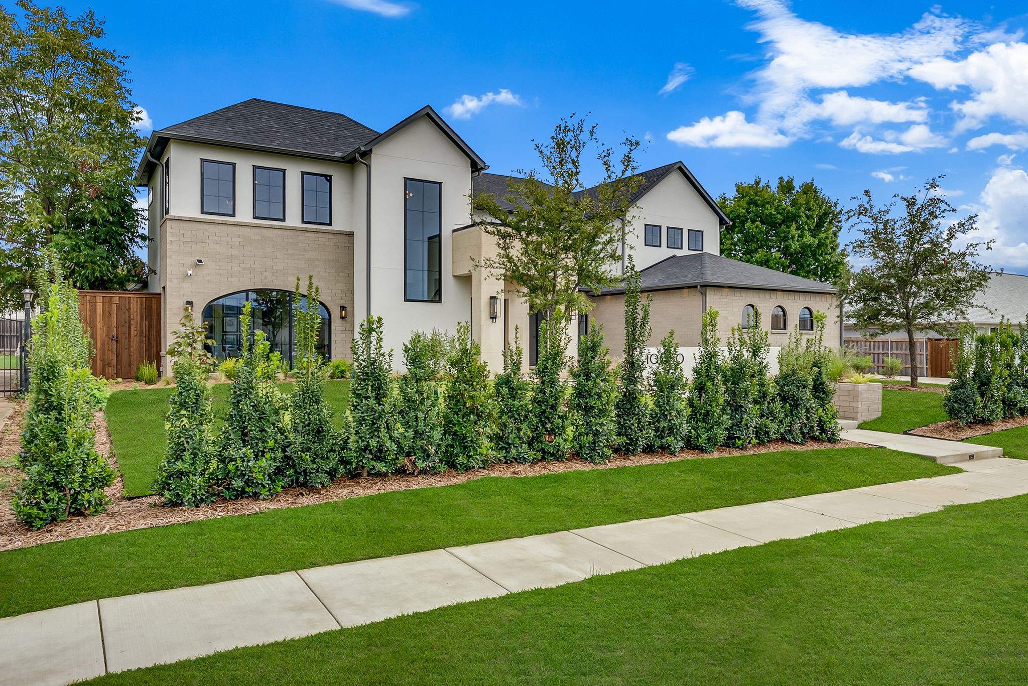 Two-story cream stucco and tan brick home with tall windows, landscaped hedges, and a broad front lawn.