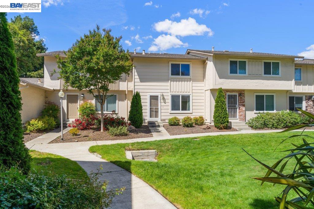 Two-story attached home with light siding, landscaped shrubs, a green lawn, and a curved front walkway.