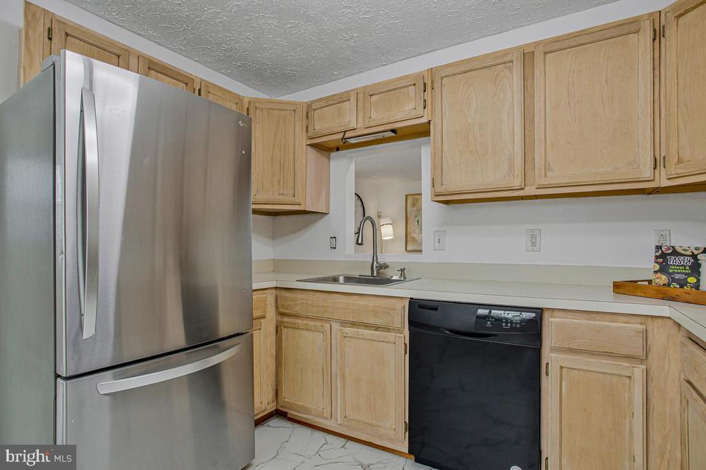 Kitchen with light wood cabinets, stainless steel refrigerator, black dishwasher, and marble-look tile flooring.