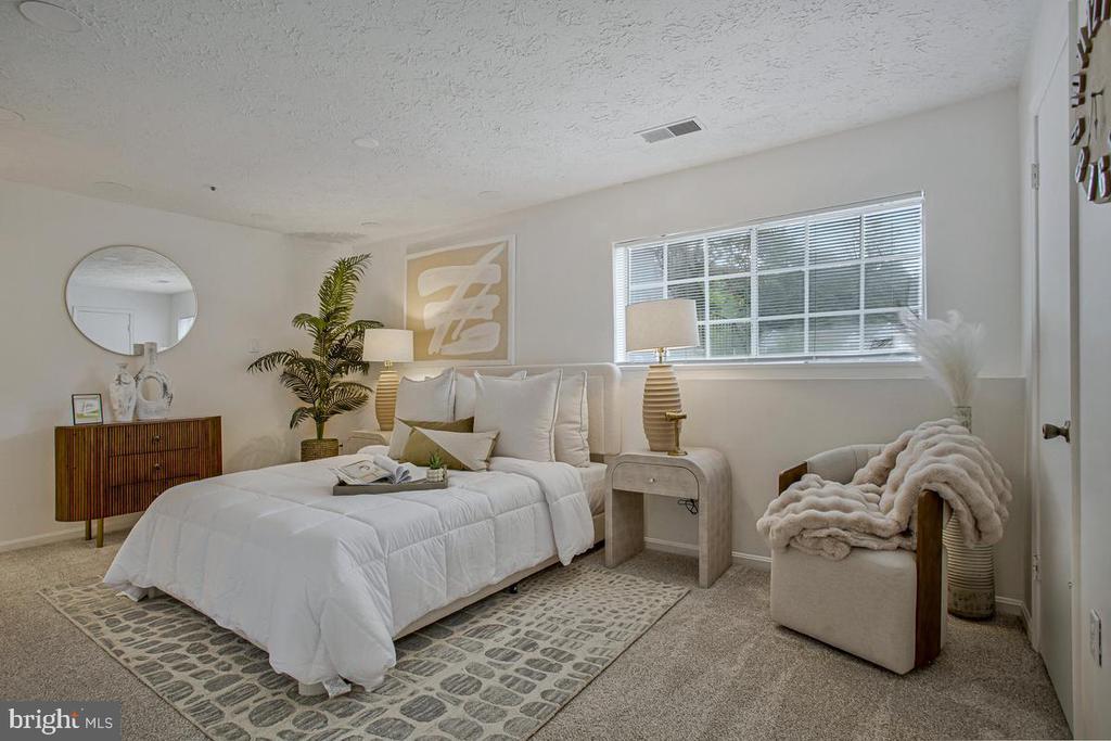 Staged bedroom with a white bed, patterned rug, wooden dresser, and large window with blinds.