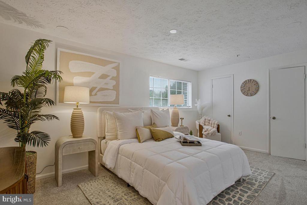 Furnished bedroom with a white quilted bed, neutral decor, and a window with blinds.