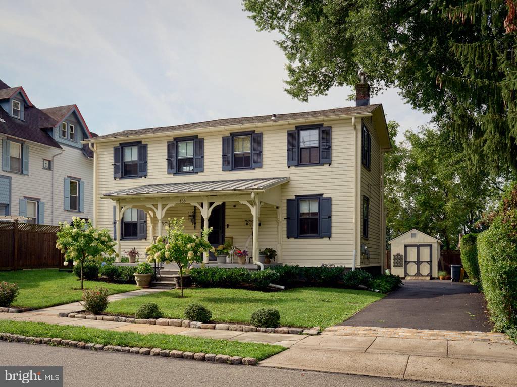 Pale yellow two-story house with dark shutters, a covered porch, and a driveway leading to a detached shed.