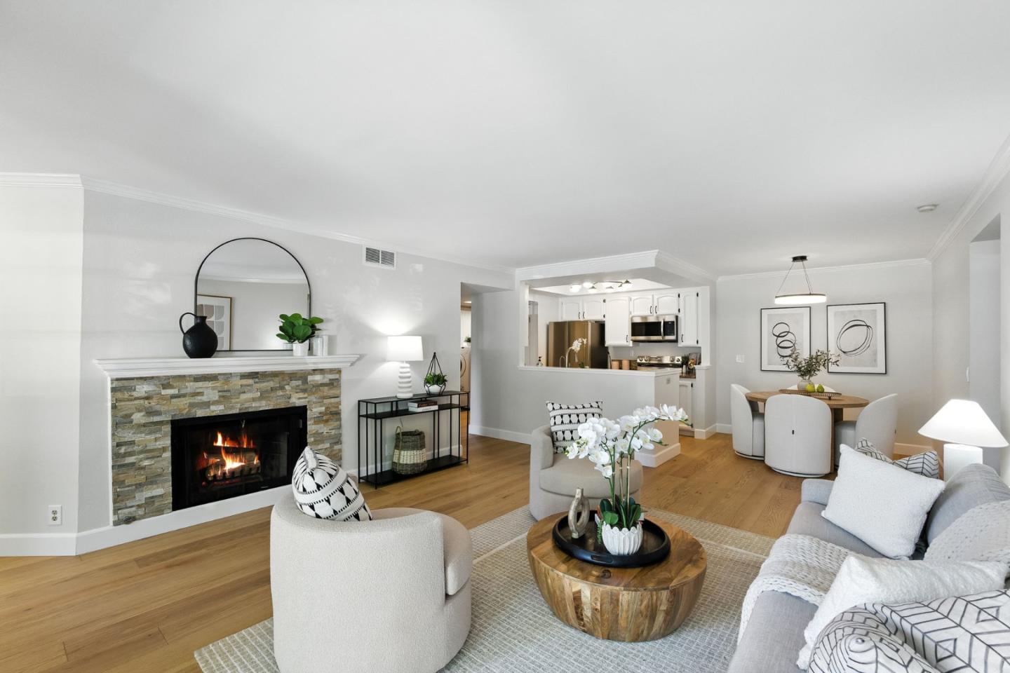 Staged living room with a stone fireplace, hardwood floors, and an open kitchen beyond.