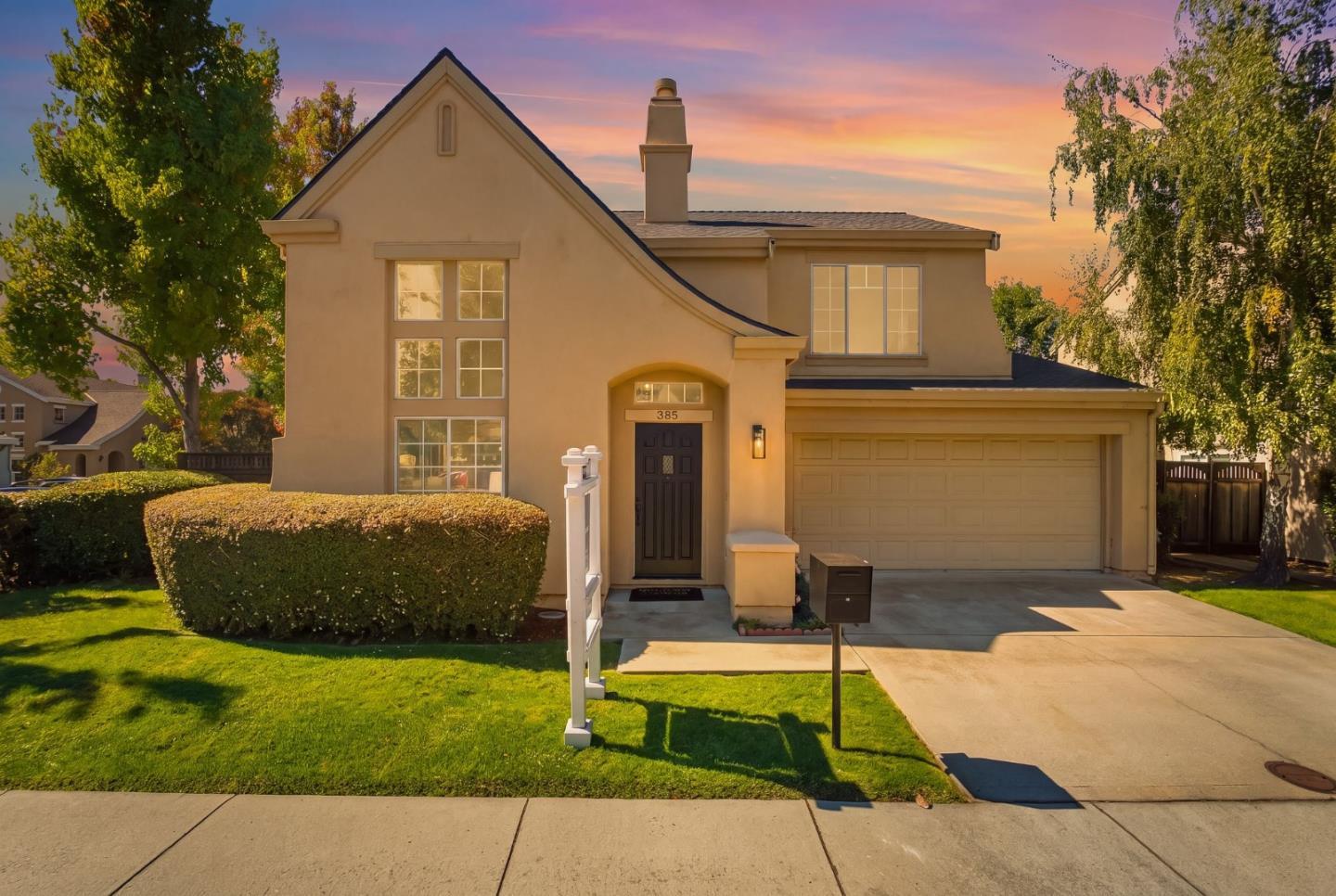 Two-story beige home with a steep gable, double garage, and sunset-lit front lawn.