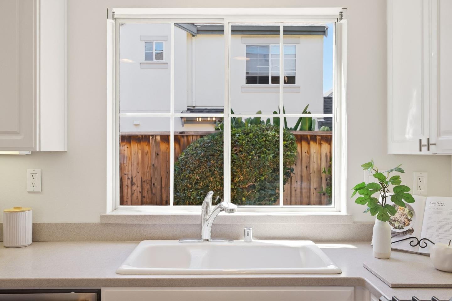 Kitchen sink beneath a large window overlooking shrubs, a wooden fence, and a neighboring house.