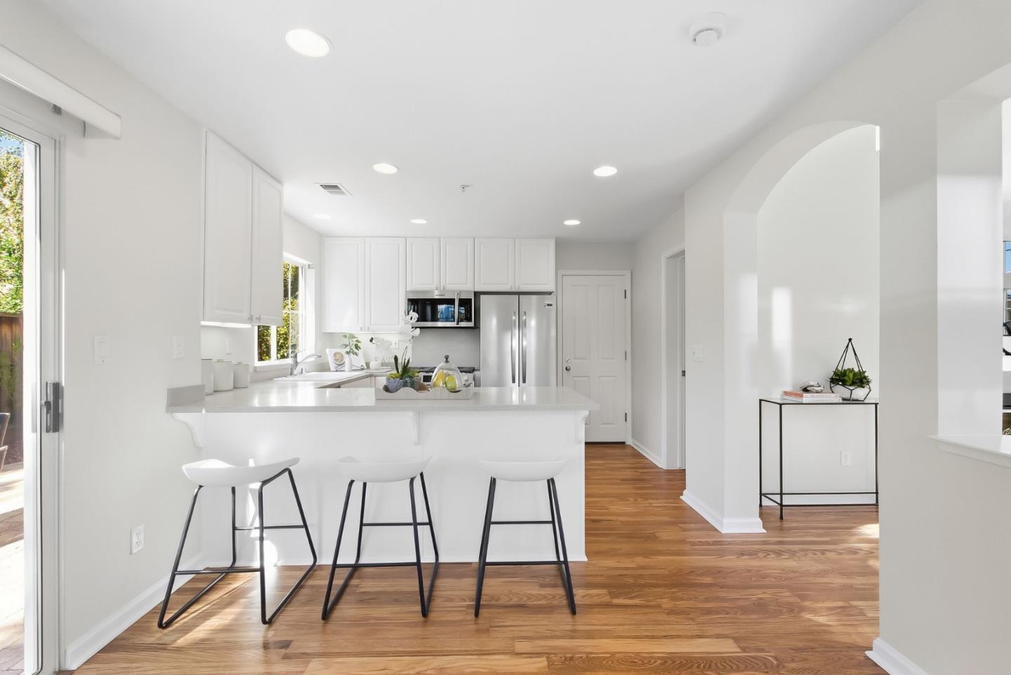 Bright white kitchen with an island breakfast bar, three stools, stainless steel appliances, and hardwood floors.