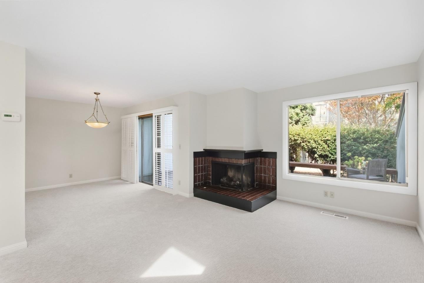 Empty living room with a tiled corner fireplace and a large window overlooking a leafy garden.