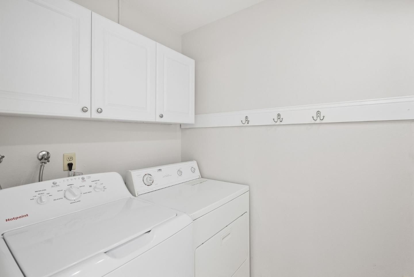 Laundry room with a white washer and dryer beneath cabinets and a row of wall hooks.