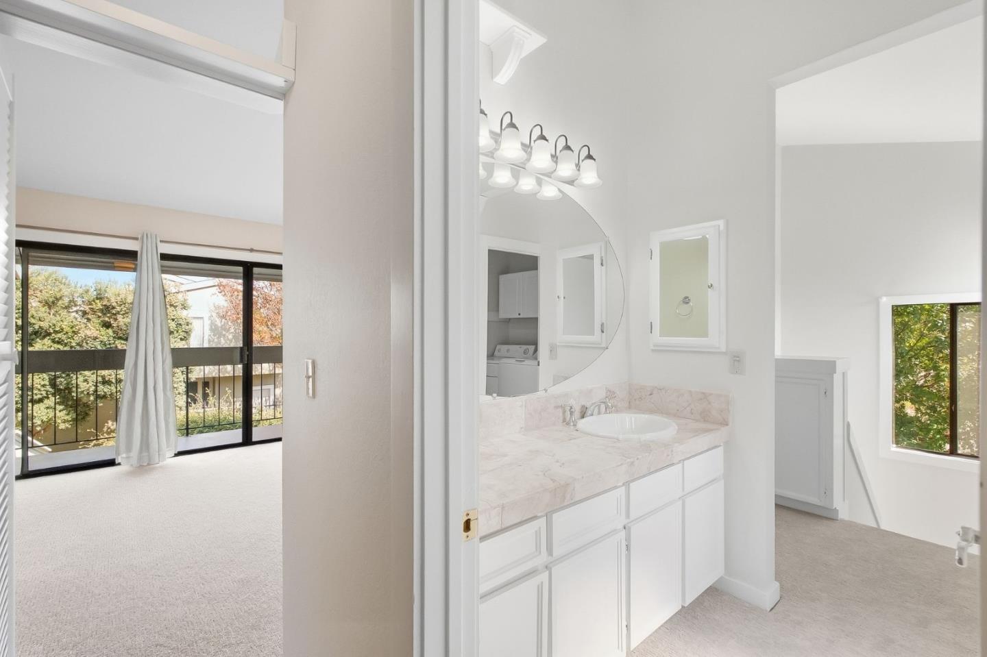 Bright bathroom vanity with a marble-pattern countertop and curved mirror beside a bedroom with sliding glass doors.