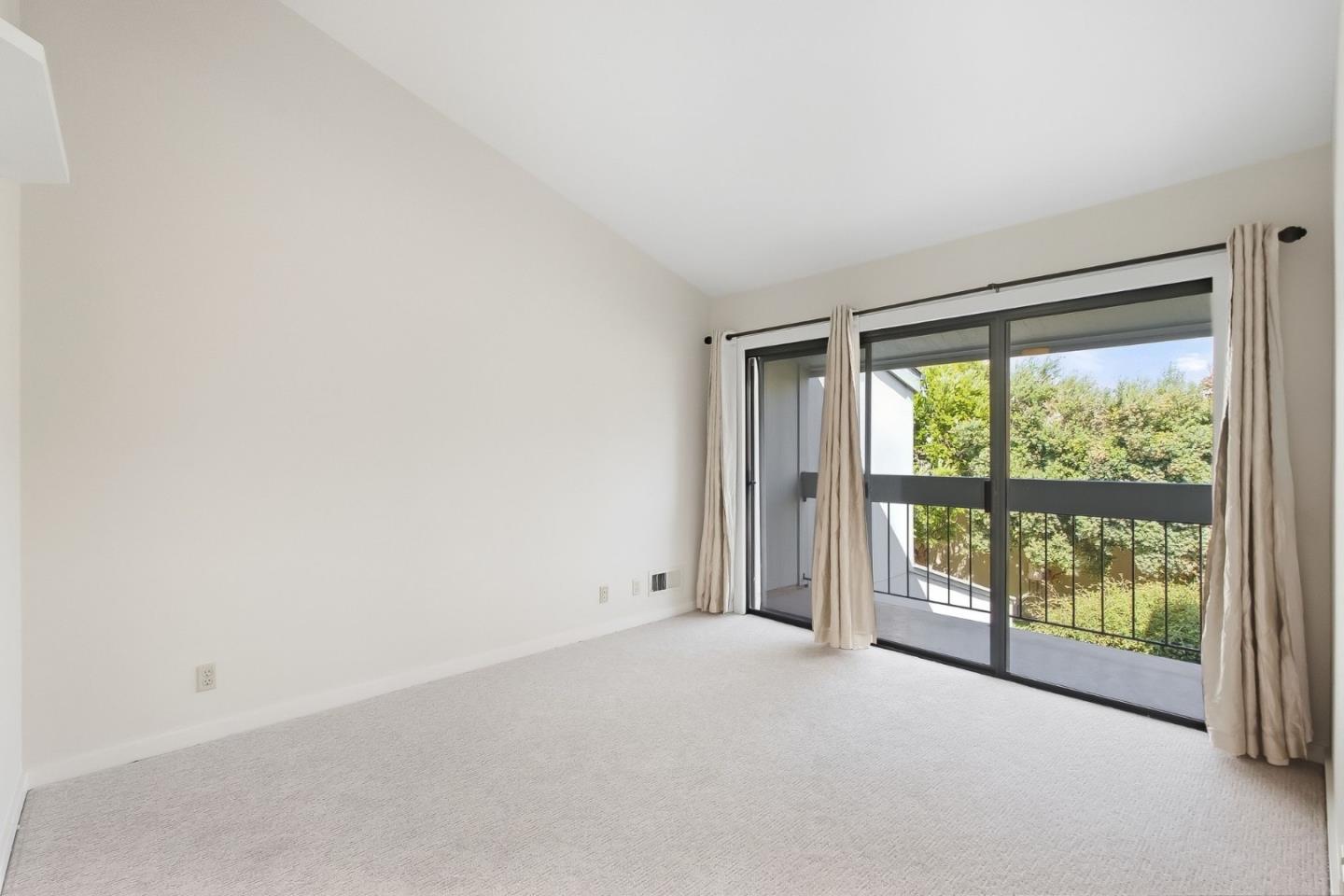 Empty carpeted room with sliding glass doors opening onto a balcony and leafy greenery.