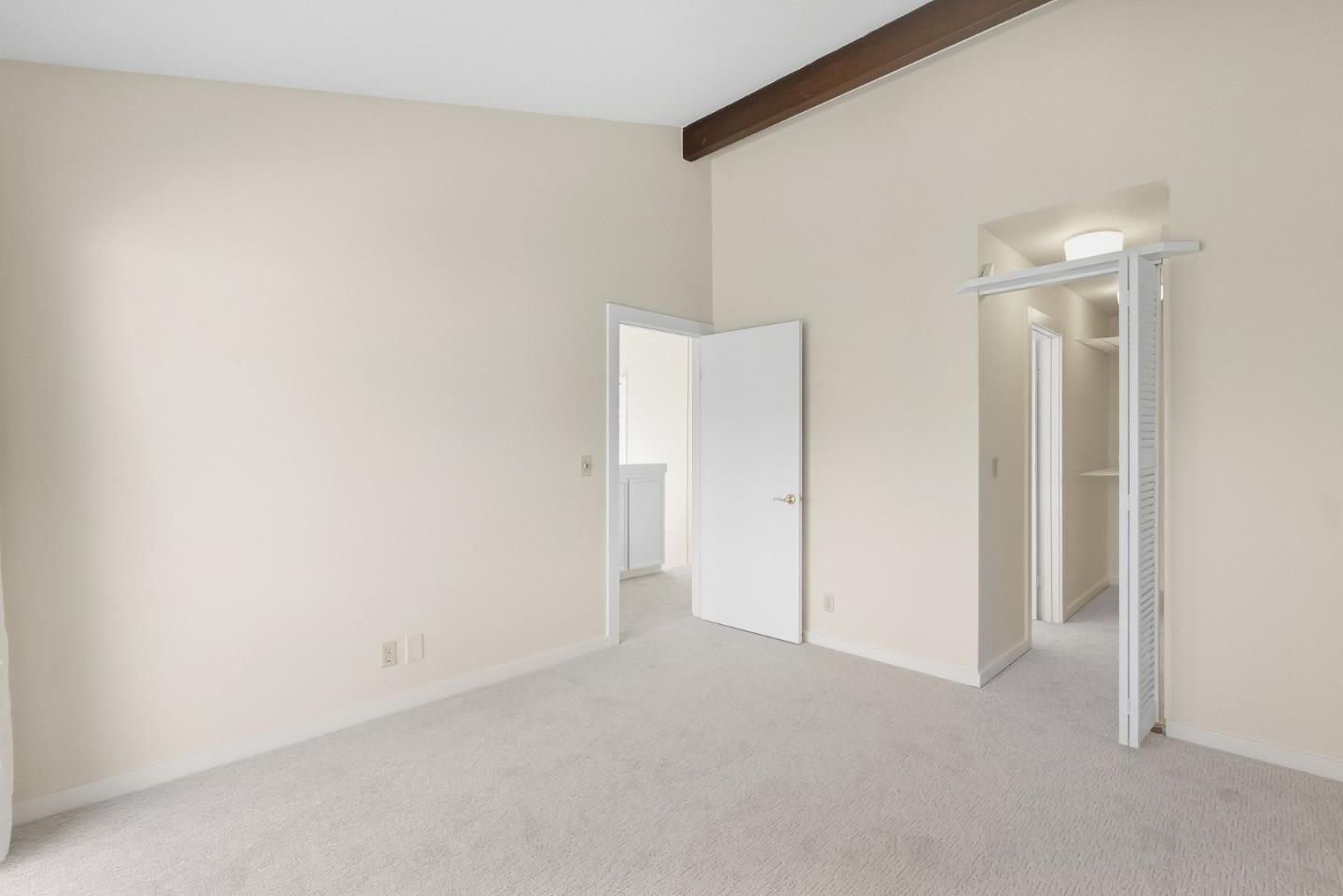 Empty bedroom with light carpet, an exposed dark ceiling beam, and an open louvered closet.