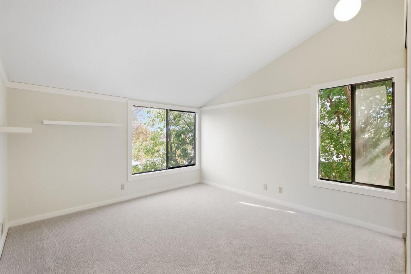 Empty carpeted room with a sloped ceiling and two large windows facing leafy trees.