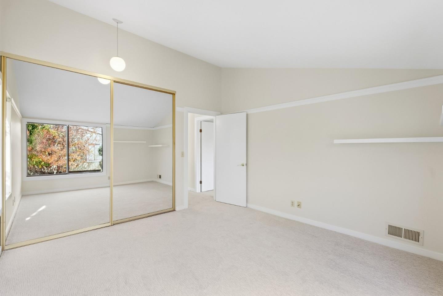 Empty bedroom with mirrored closet doors, light carpet, and a window overlooking autumn foliage.