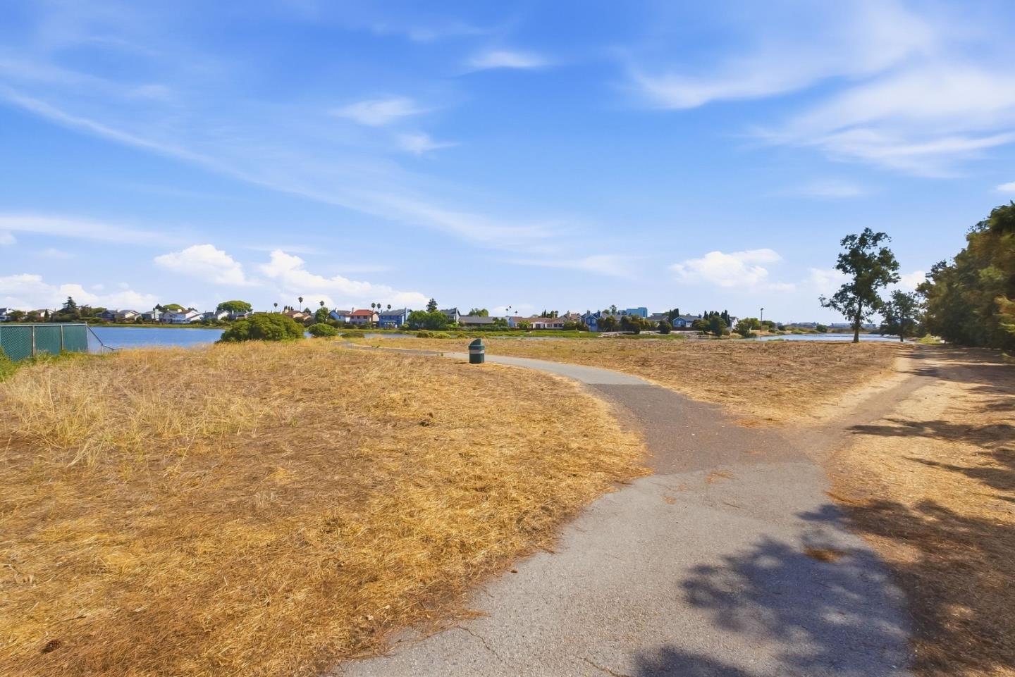 A paved path curves through a dry lakeside park, with homes across the water.