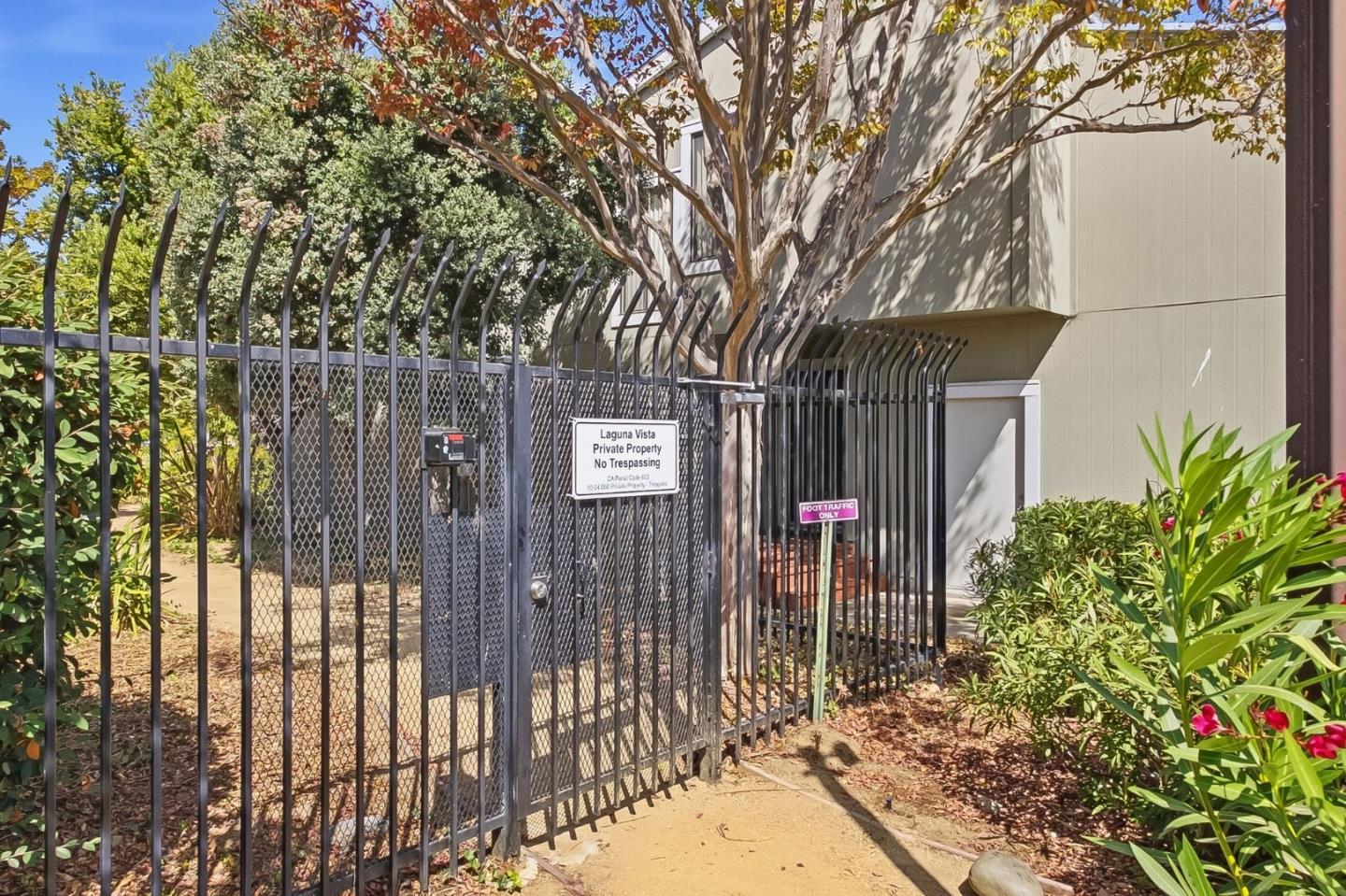 Gated residential entrance with tall black metal fencing, a pedestrian gate, and mature trees.