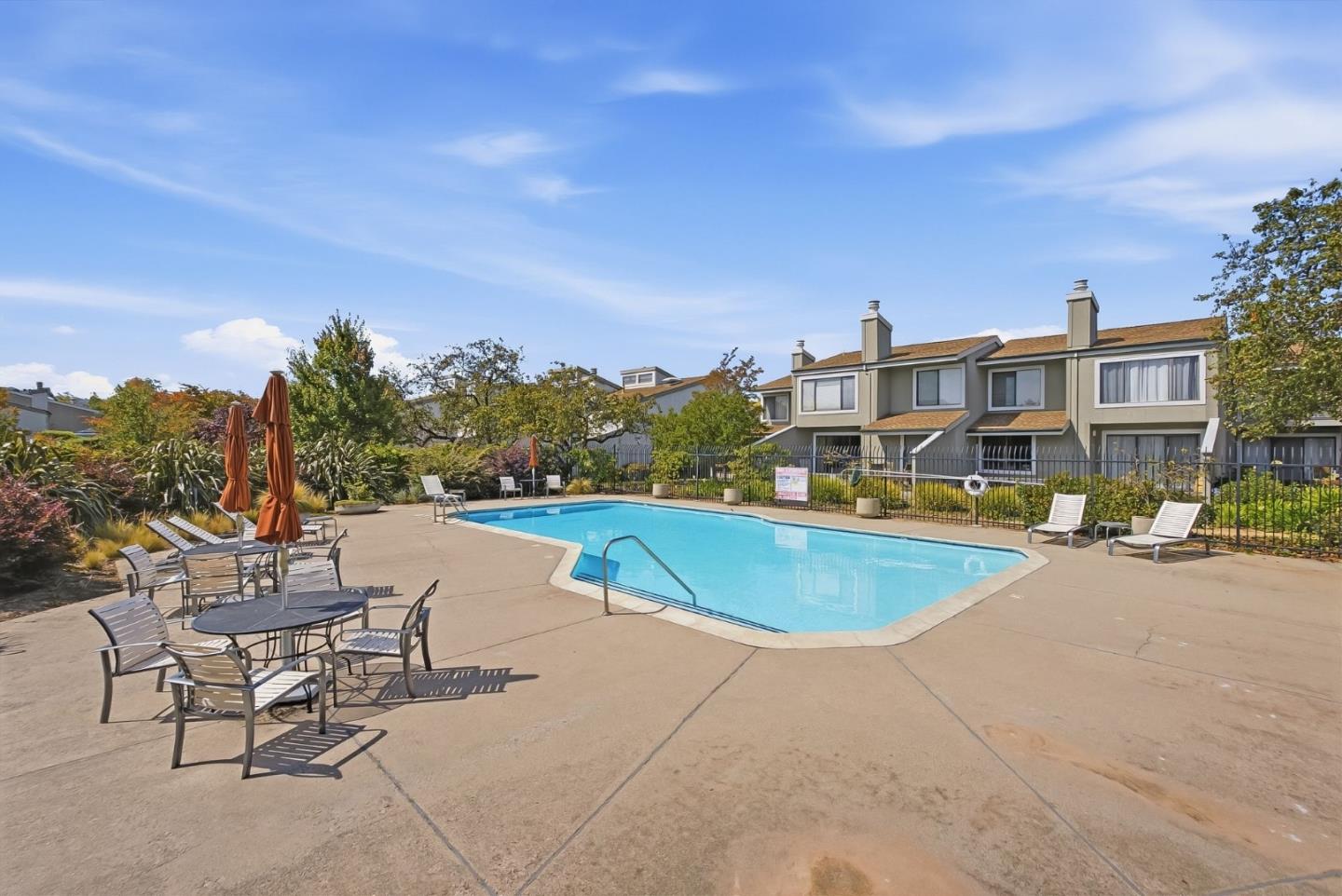 Outdoor pool beside attached homes, with patio tables, lounge chairs, and landscaped grounds.