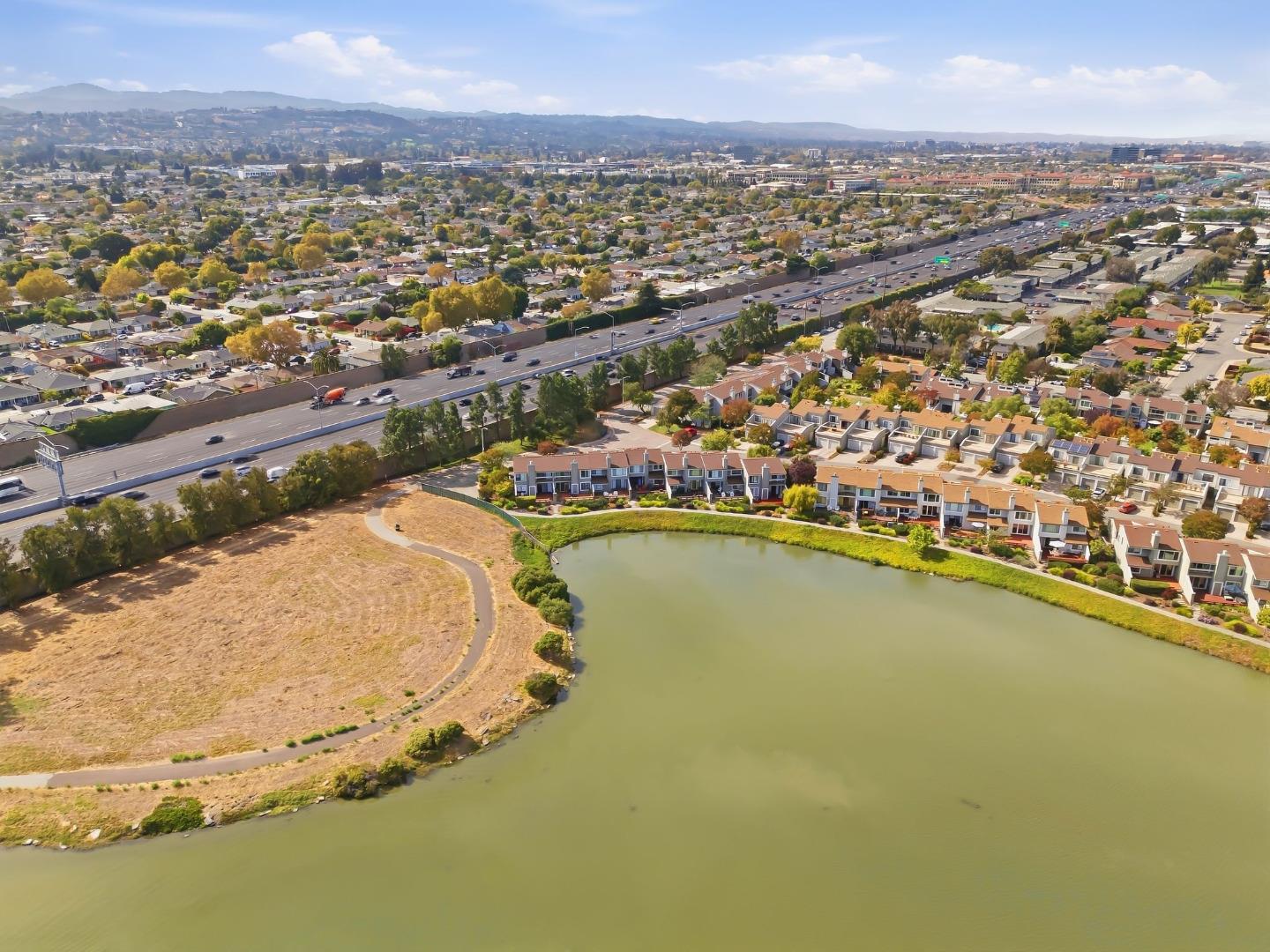 Aerial view of homes beside a pond, with a walking path, highway, and distant hills.