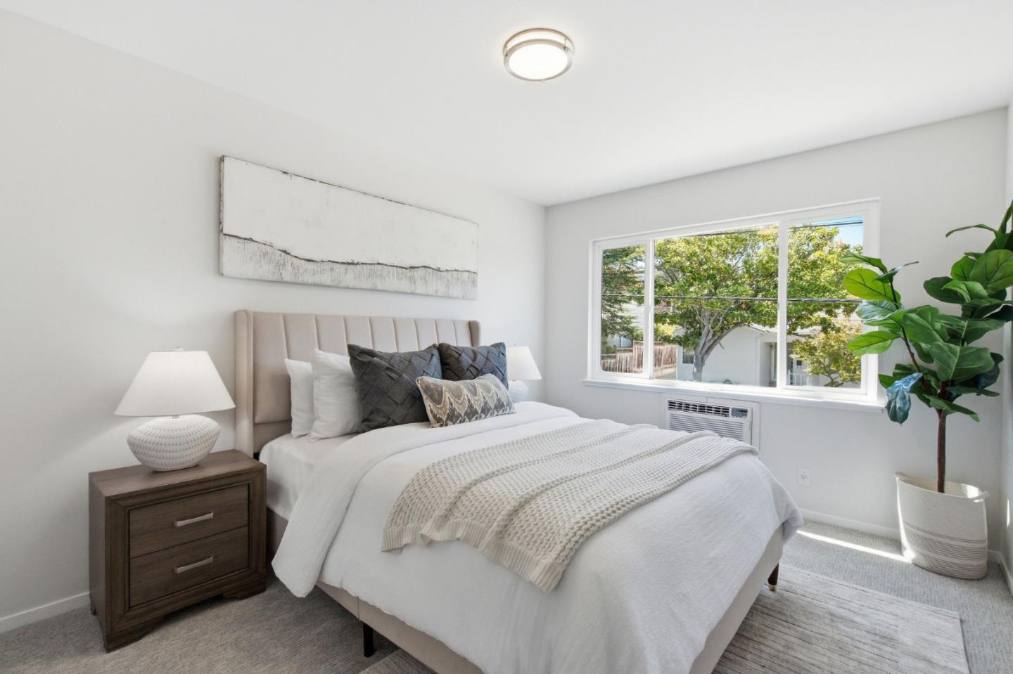 Staged bedroom with a large white bed, upholstered headboard, and wide window overlooking trees.