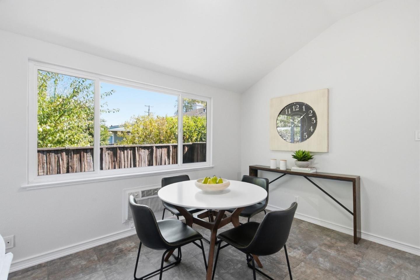 Dining area with a round table and four chairs beside a large window overlooking a fenced yard.