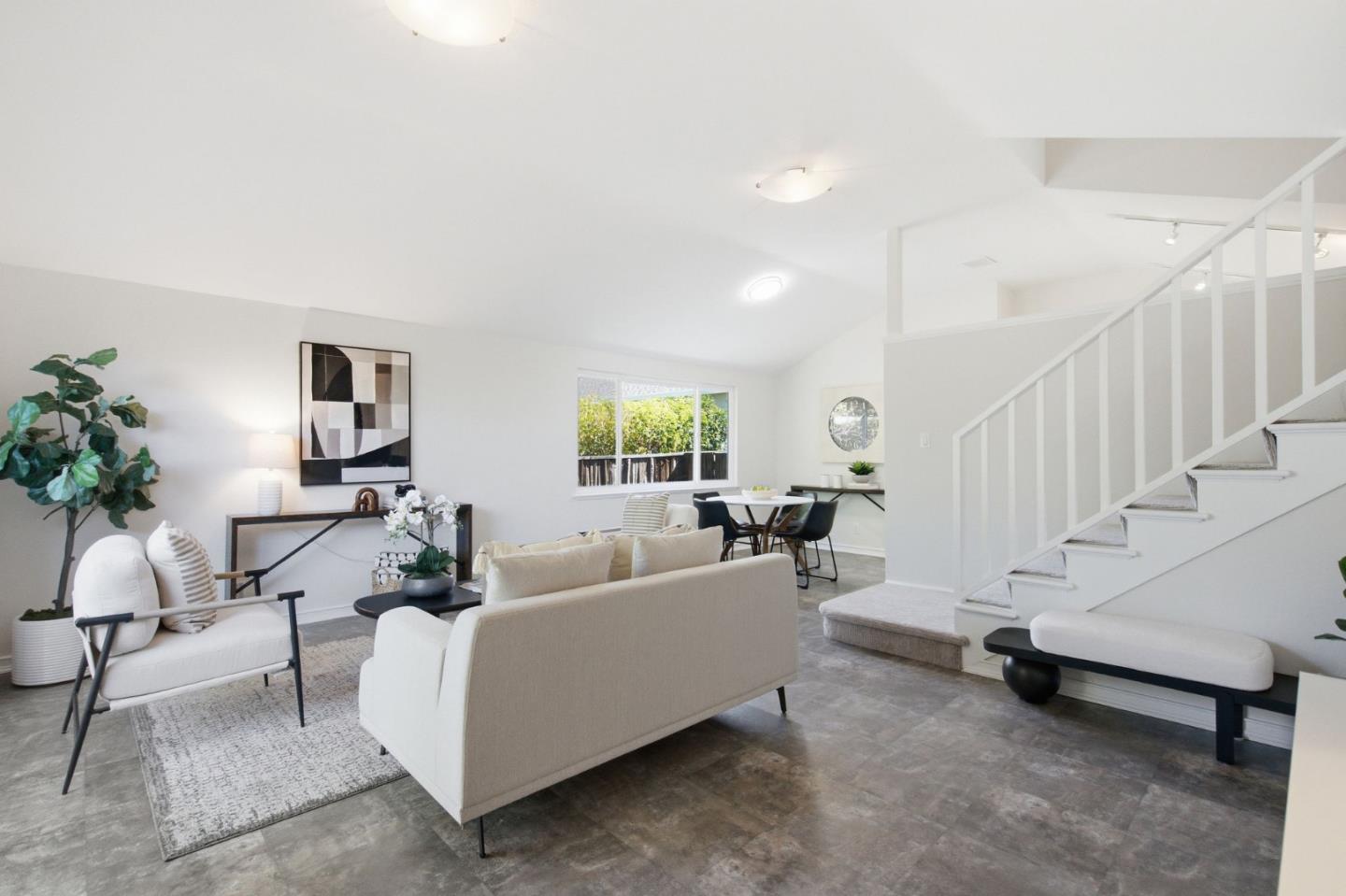Staged living room with neutral seating, gray tile floors, and a staircase beside the dining area.