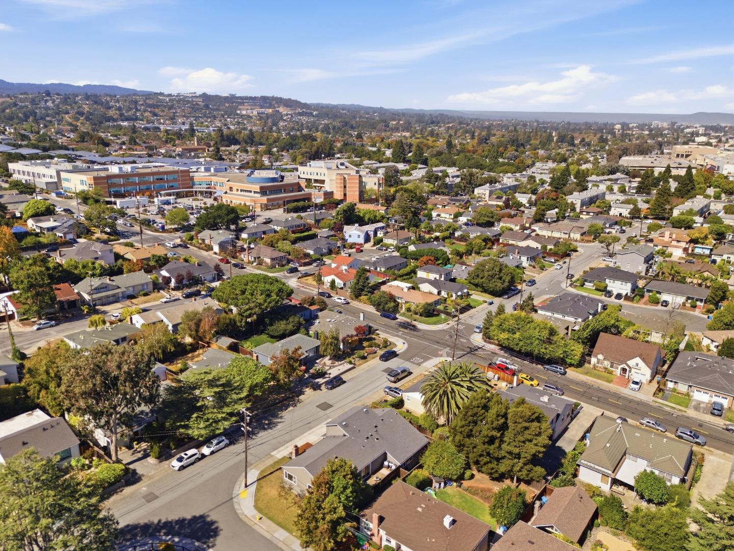Aerial view of San Mateo homes and streets, with commercial buildings and low hills in the distance.