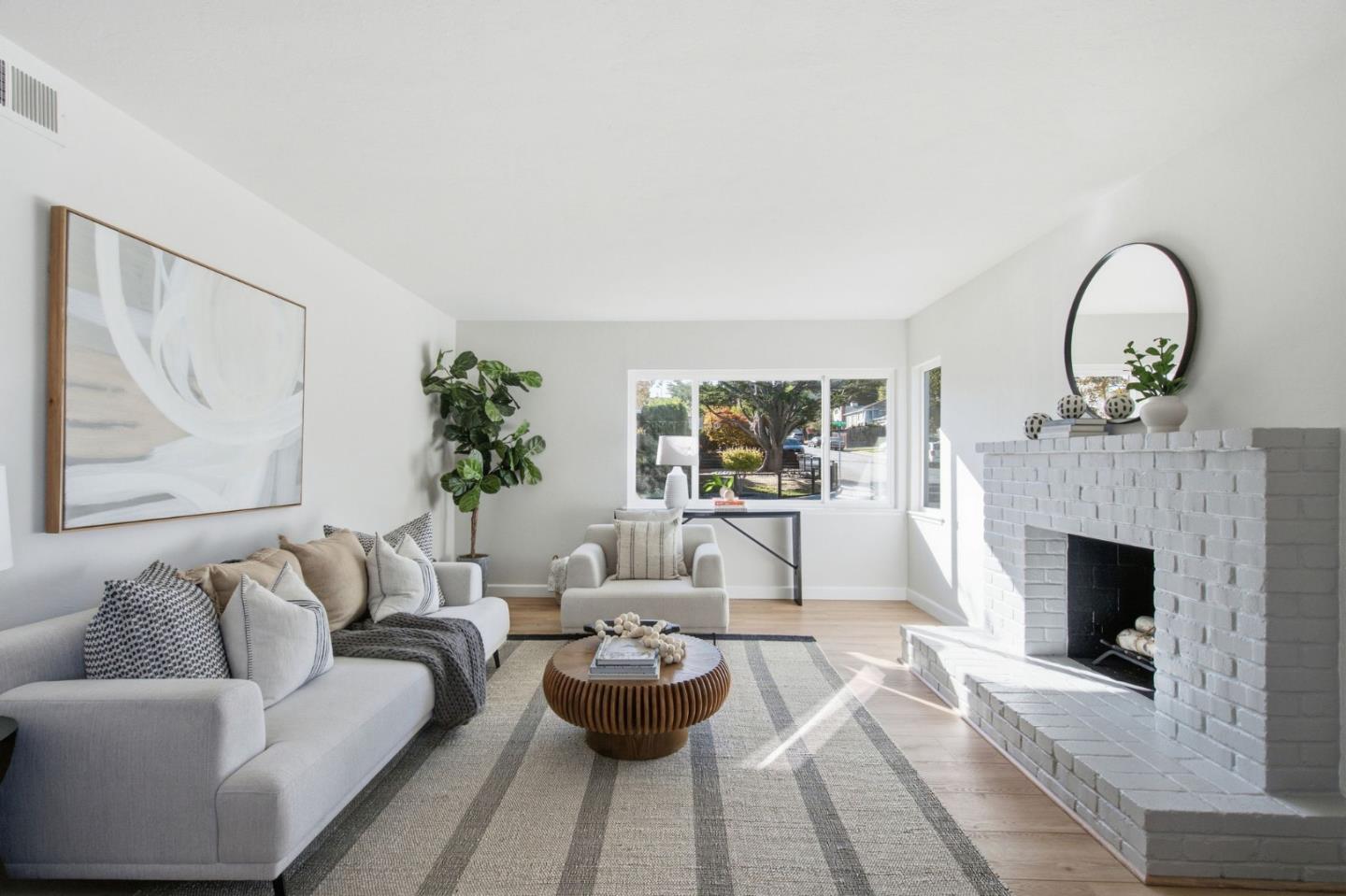 Staged living room with a white brick fireplace, gray seating, and sunlight streaming through large windows.