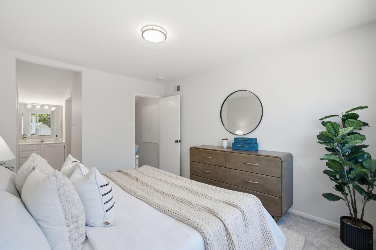 Staged bedroom with a white bed, wood dresser, round mirror, and potted plant.