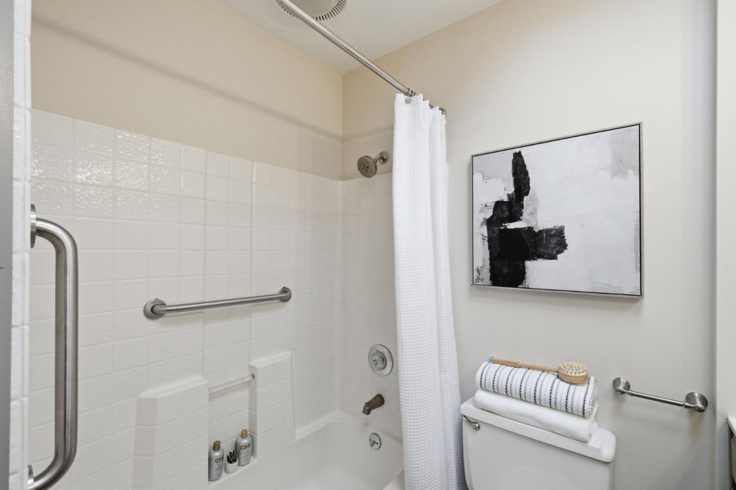 Bathroom with a white tiled tub, shower curtain, grab bars, and abstract wall art.