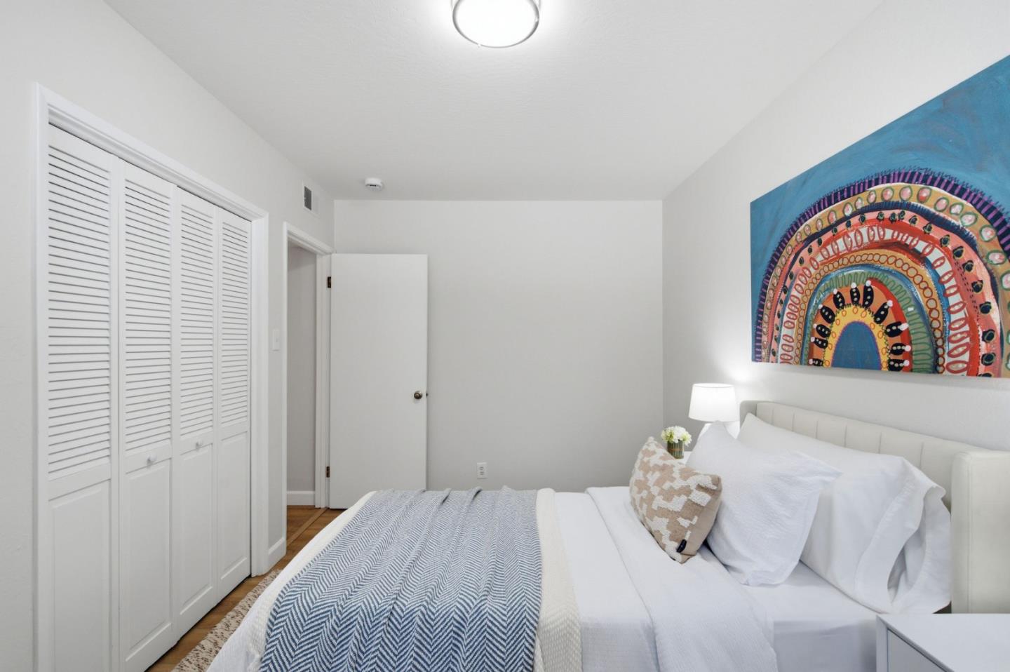 Bedroom with white bedding, louvered closet doors, and colorful semicircular wall art.
