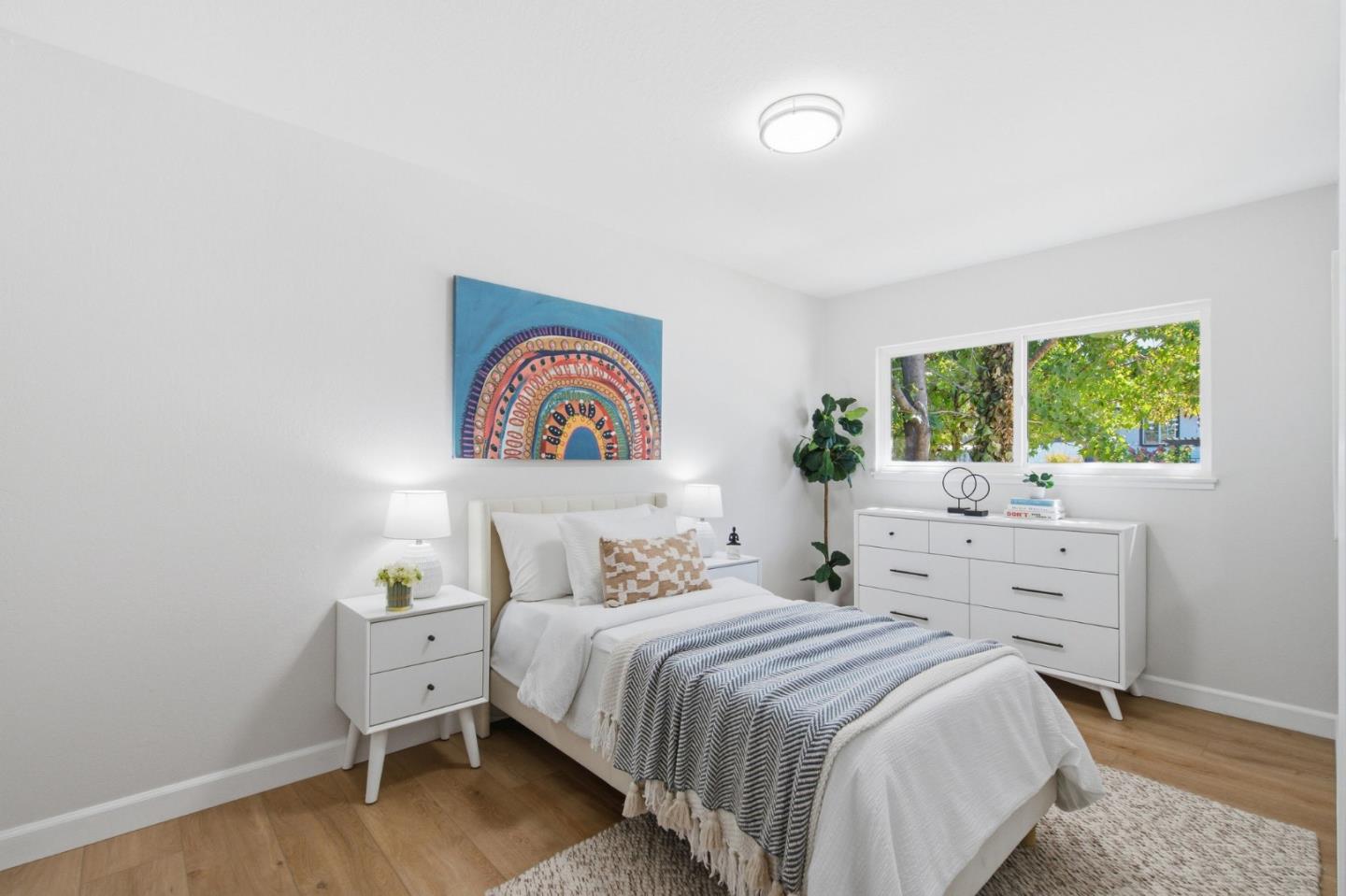 Bright bedroom with a white bed, striped throw, colorful wall art, and a window overlooking trees.