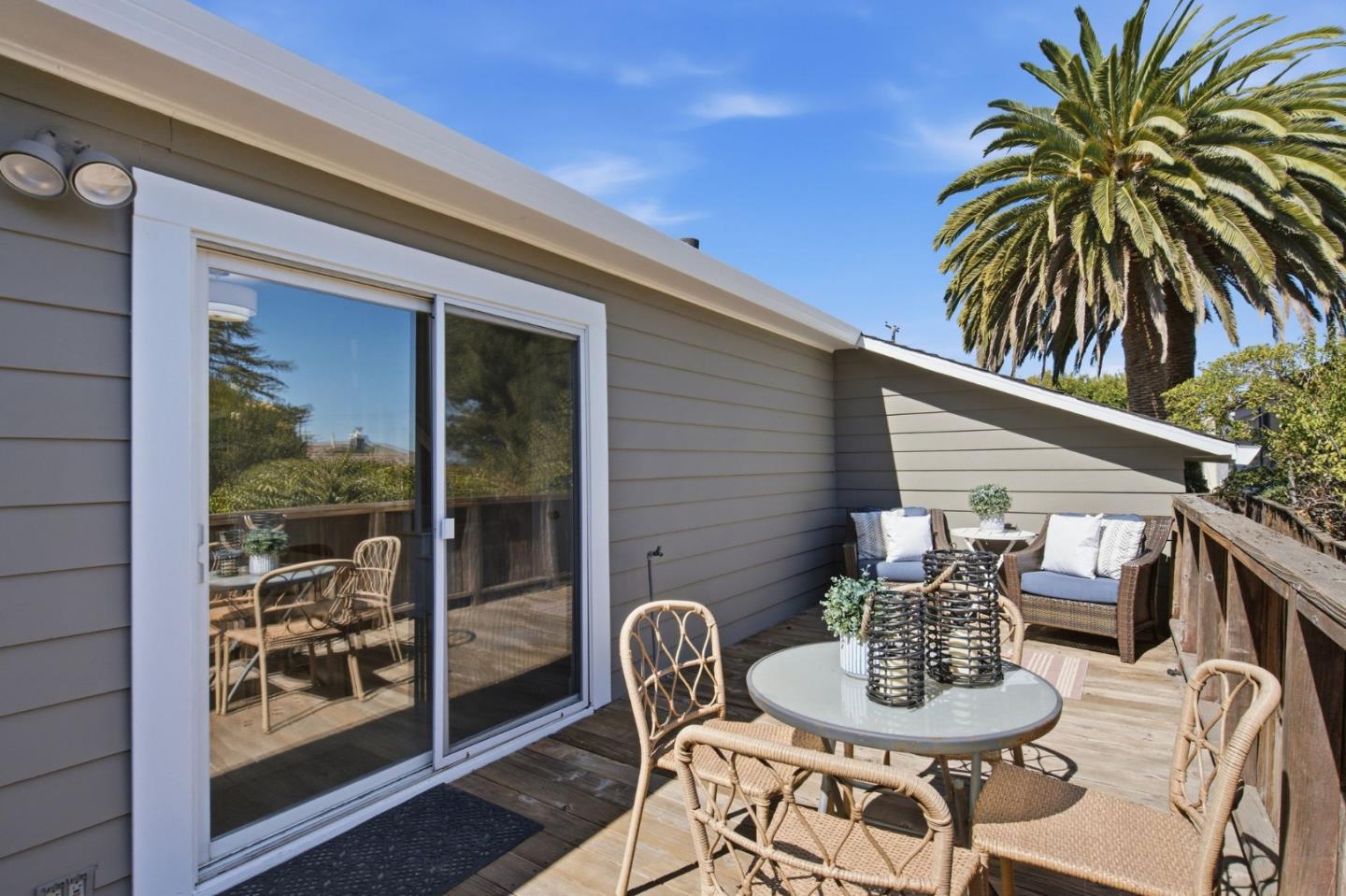 Wooden deck with wicker seating and a round table beside a gray-sided home, with a tall palm tree.