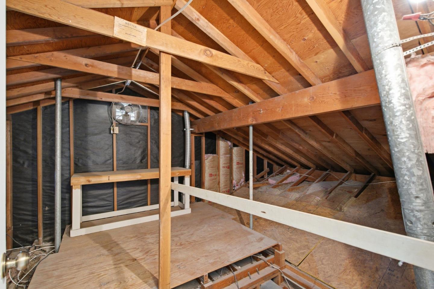 Unfinished attic with exposed wooden trusses, insulation, and a metal ventilation duct.
