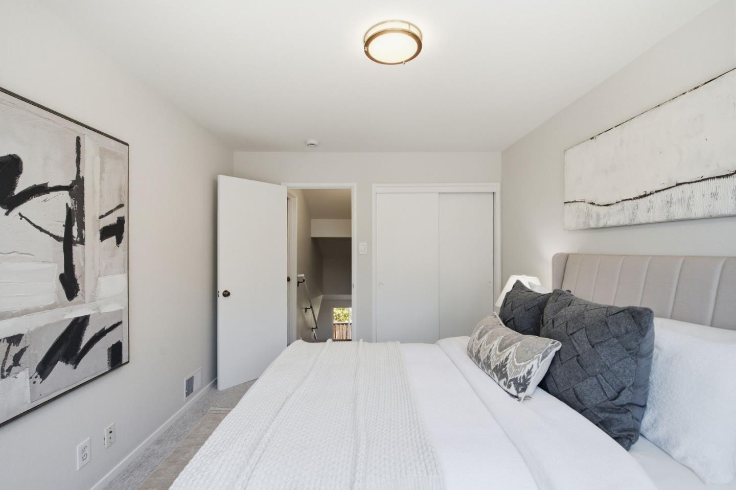 Staged bedroom with a white bed, gray pillows, abstract art, and an open doorway to the stairs.