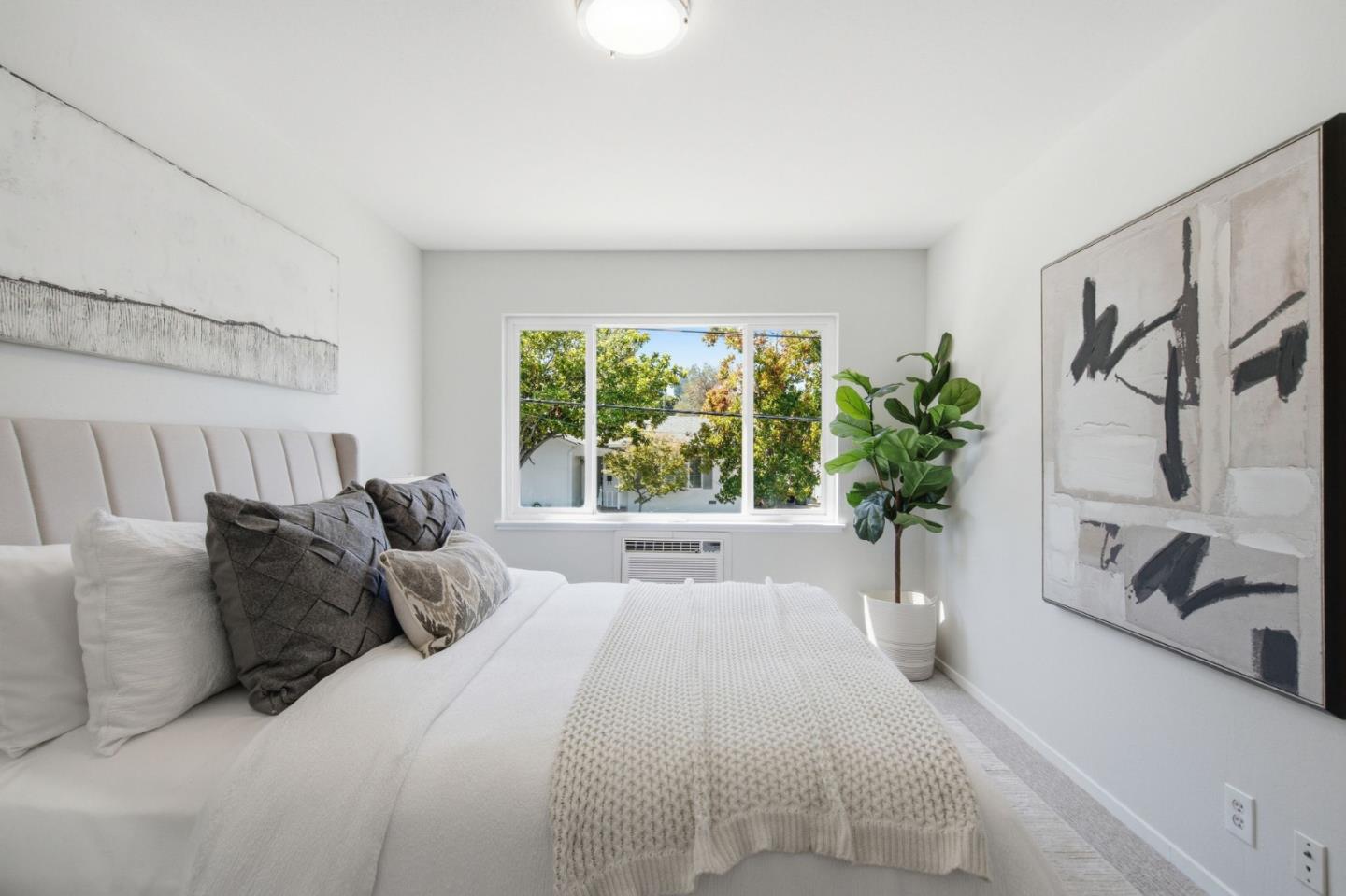 Bright staged bedroom with a white upholstered bed, wide window, abstract art, and potted plant.