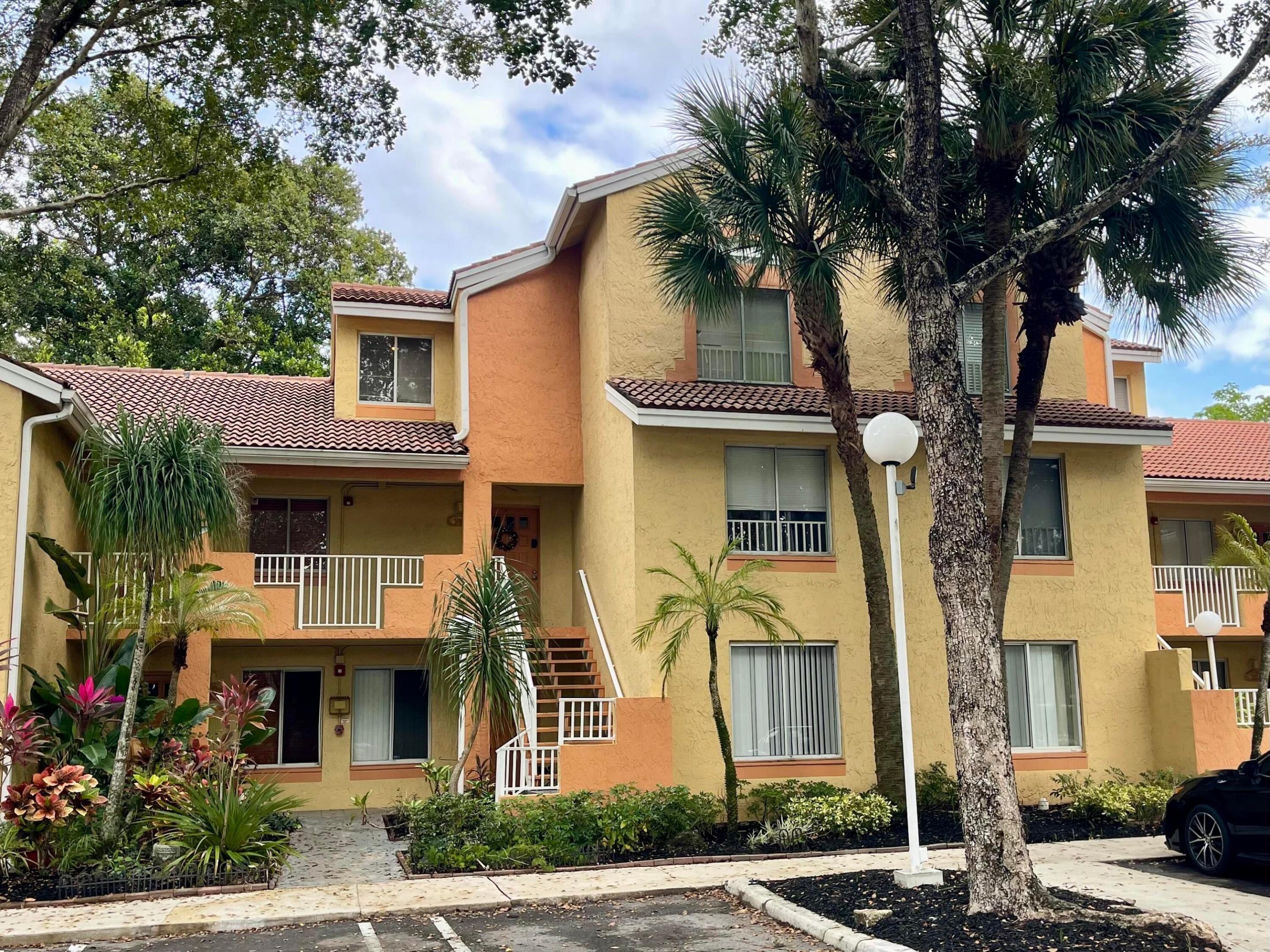 Two-story yellow and coral condominium building with red tile roofs, balconies, and palm trees.