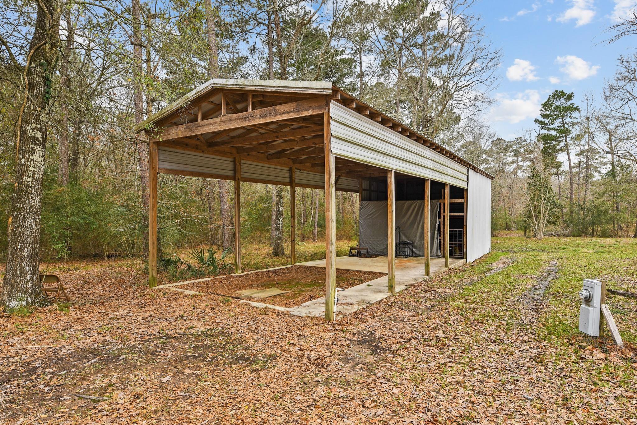 Open-sided pole barn with white metal siding and exposed wooden supports in a wooded clearing.